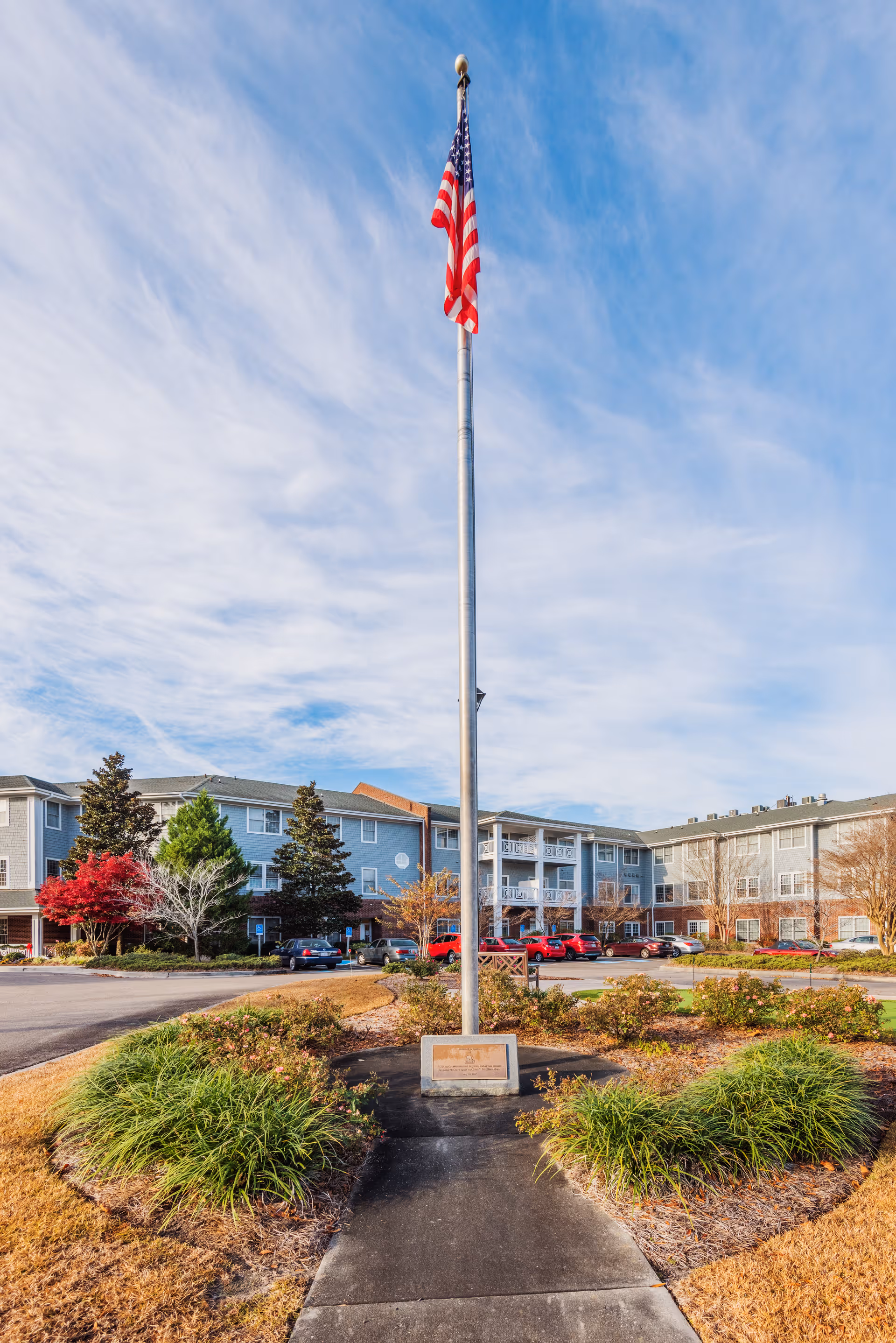 View of the exterior of Brightmore of Wilmington senior living facility with a tall flagpole flying the American flag in the foreground, surrounded by landscaped bushes and a paved walkway leading to the flagpole. The multi-story building with balconies and parked cars is visible in the background under a partly cloudy sky.