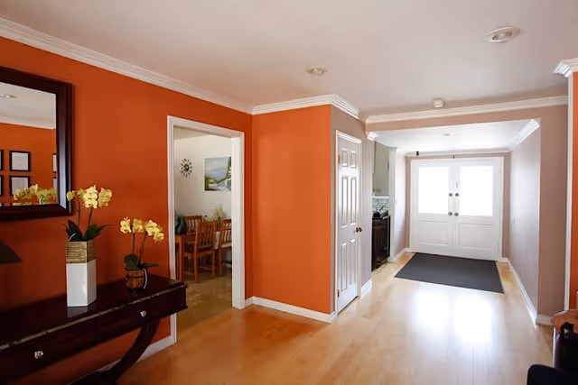 Interior hallway with orange walls and light wood flooring leading to a set of white double doors. A dark wooden console table with yellow orchids in white and brown pots is on the left side under a large mirror. Through an open doorway on the left, a dining area with a wooden table and chairs is visible.