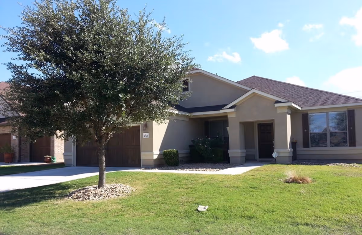 Single-story residential building with beige exterior walls and a dark brown roof. There is a large tree in the front yard, a driveway leading to a double garage with wooden doors, and a small covered porch at the entrance. The lawn is green and well-maintained under a partly cloudy blue sky.