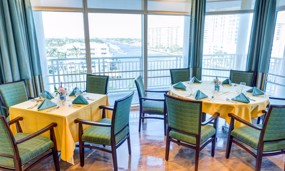 Sunlit dining room with round and square tables set with yellow tablecloths and green napkins overlooking a waterfront through large windows.