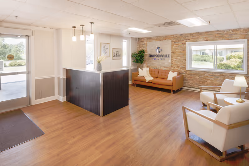 Reception area of Simpsonville Post-Acute facility featuring a dark wood reception desk with a vase of flowers, a brown leather couch with pillows against a brick accent wall, two white armchairs with wooden frames, a small round table with a lamp, large windows letting in natural light, and a glass door entrance.