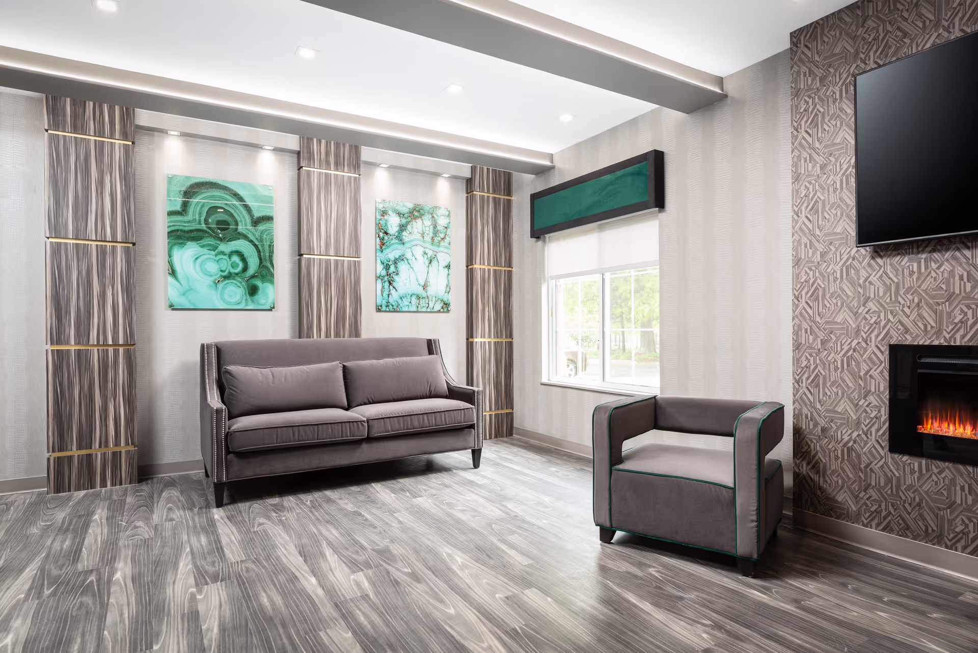 A modern living room with a gray sofa and matching armchair, wood-patterned flooring, two green abstract paintings on the wall, a window with a green valance, and a wall-mounted flat screen TV above an electric fireplace.
