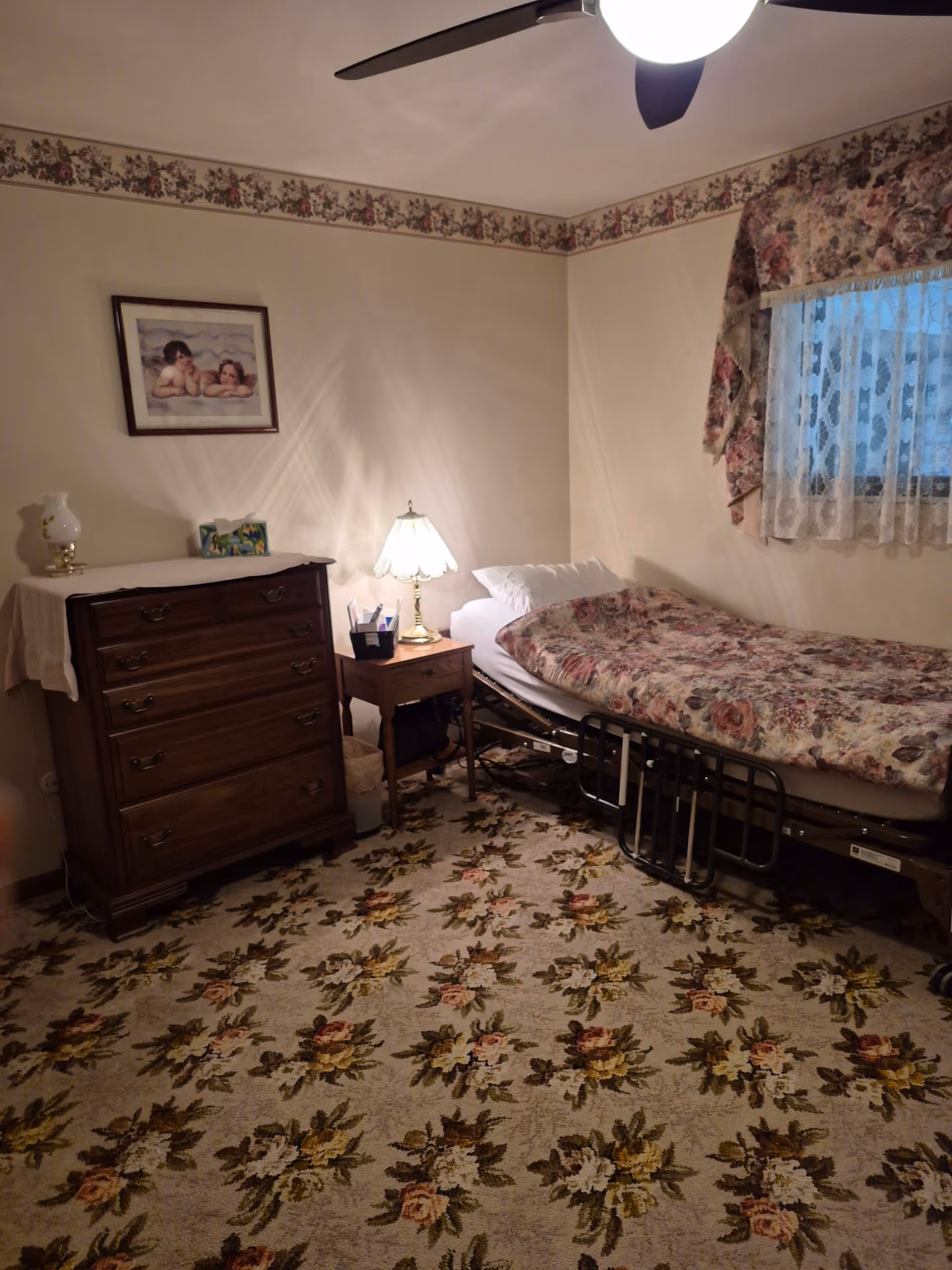 A small bedroom with a single bed covered in floral bedding, a wooden nightstand with a lamp, a wooden dresser with a white cloth and decorative items on top, floral carpet, floral valance and lace curtains on the window, and a ceiling fan with a light.