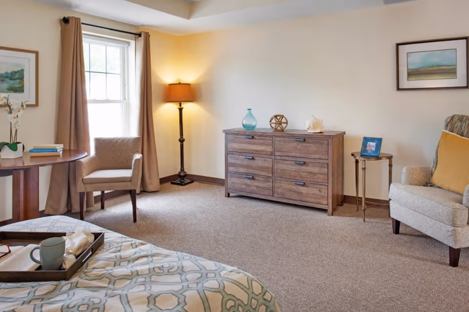 A cozy bedroom in a senior living facility with a bed partially visible in the foreground, a wooden dresser against the wall with decorative items on top, a floor lamp in the corner, an armchair with a yellow cushion, a small round side table with a framed photo, and a window with beige curtains letting in natural light.