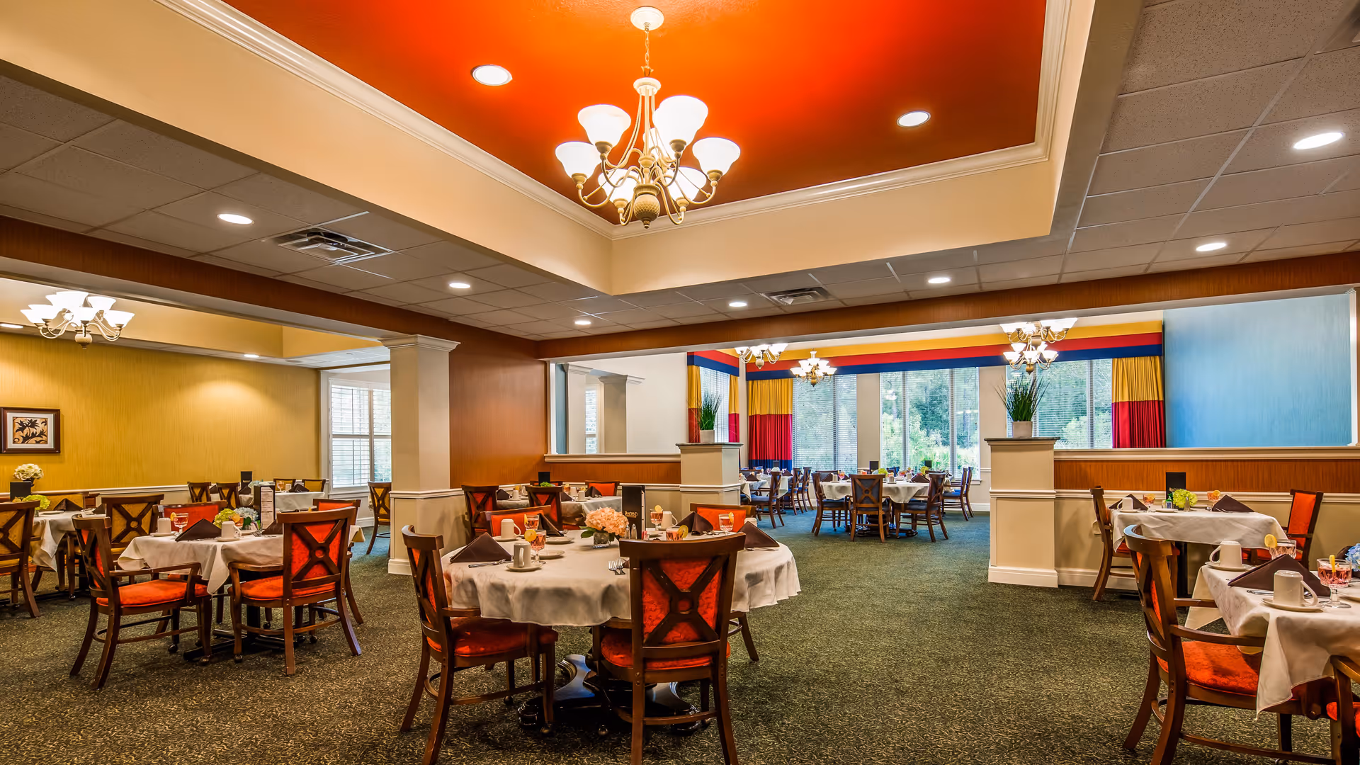 A spacious dining room with multiple round tables covered with white tablecloths, each set with cups, napkins, and small flower arrangements. The room features warm lighting with chandeliers, a red ceiling accent, and large windows with colorful curtains allowing natural light to enter.