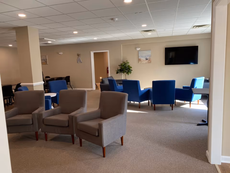 A spacious common area in an assisted living facility featuring multiple seating arrangements with gray and blue armchairs. The room has beige walls, a carpeted floor, a flat-screen TV mounted on the wall, a potted plant, and ceiling lights providing bright illumination.
