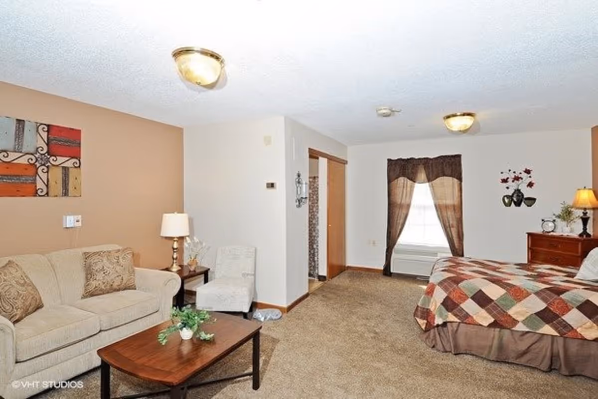 A cozy room with a beige sofa and patterned pillows on the left, a wooden coffee table with a small plant, a beige armchair, and a bed with a multicolored patchwork quilt on the right. The room has beige carpet, a window with brown curtains, a wooden dresser with a lamp and decorative items, and a wall with a piece of colorful abstract art.