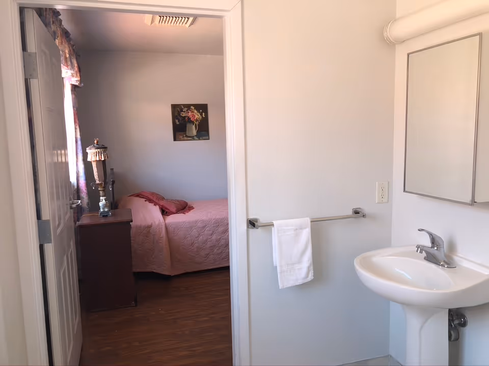 View of a small bedroom with a pink bedspread and pillows, a wooden nightstand with a decorative lamp, and a floral painting on the wall. The bedroom is seen through an open door from an adjacent bathroom area that has a white pedestal sink, a mirror cabinet, and a towel hanging on a metal bar.