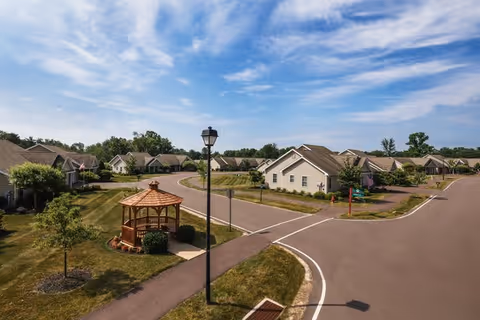 A peaceful residential community with single-story houses arranged along curved roads under a partly cloudy blue sky. There is a wooden gazebo surrounded by greenery and a streetlamp near the center of the image.