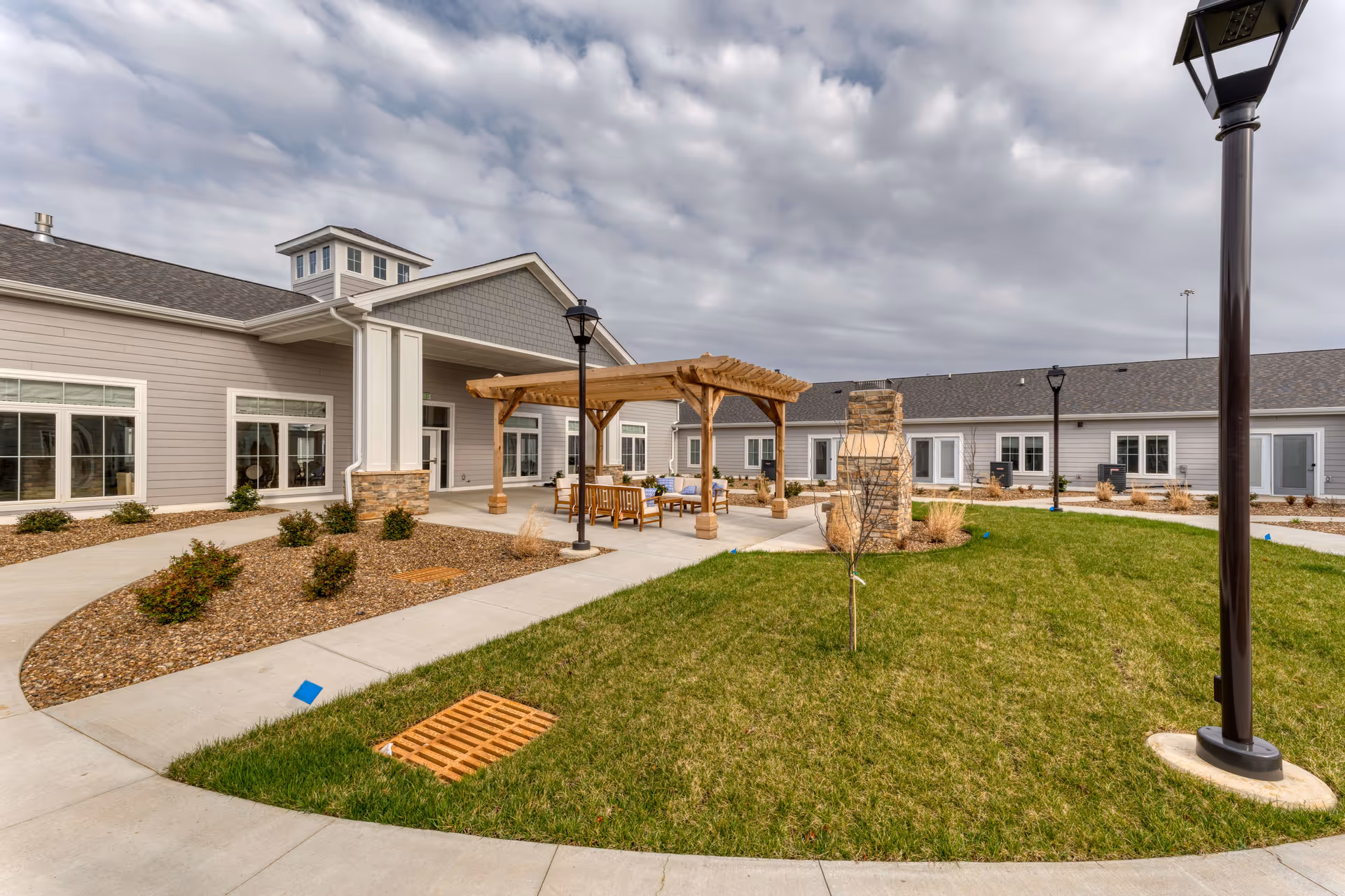 Outdoor courtyard area at Cedarhurst Senior Living of Owensboro featuring a wooden pergola with seating underneath, a stone fireplace, green lawn, landscaped bushes, concrete walkways, and lamp posts under a cloudy sky.