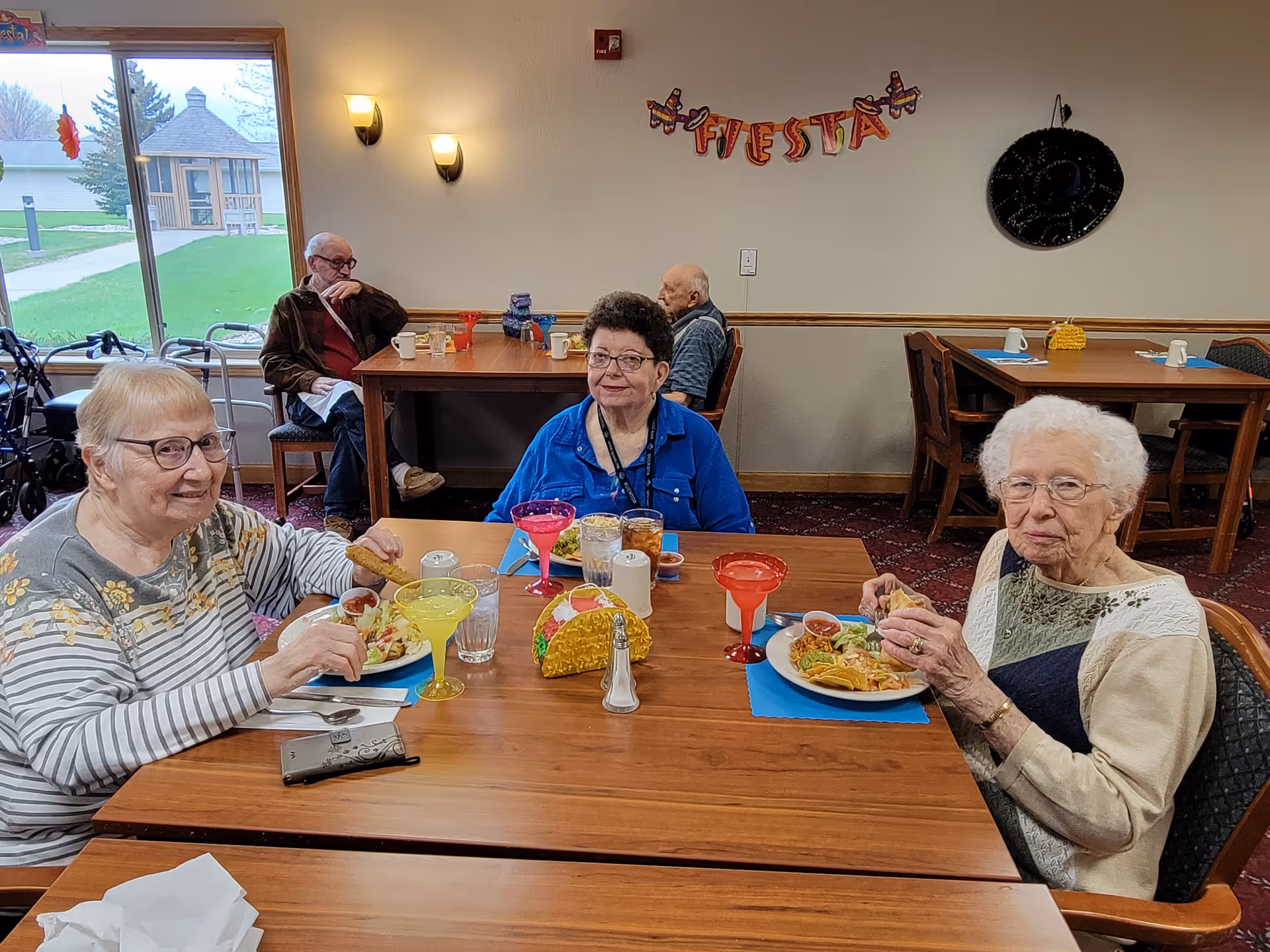 Three elderly women sitting at a dining table in a senior living facility enjoying a meal with tacos and colorful drinks. Behind them, two elderly men are seated at another table near a window showing an outdoor gazebo. The wall has a festive 'FIESTA' banner and a decorative sombrero.