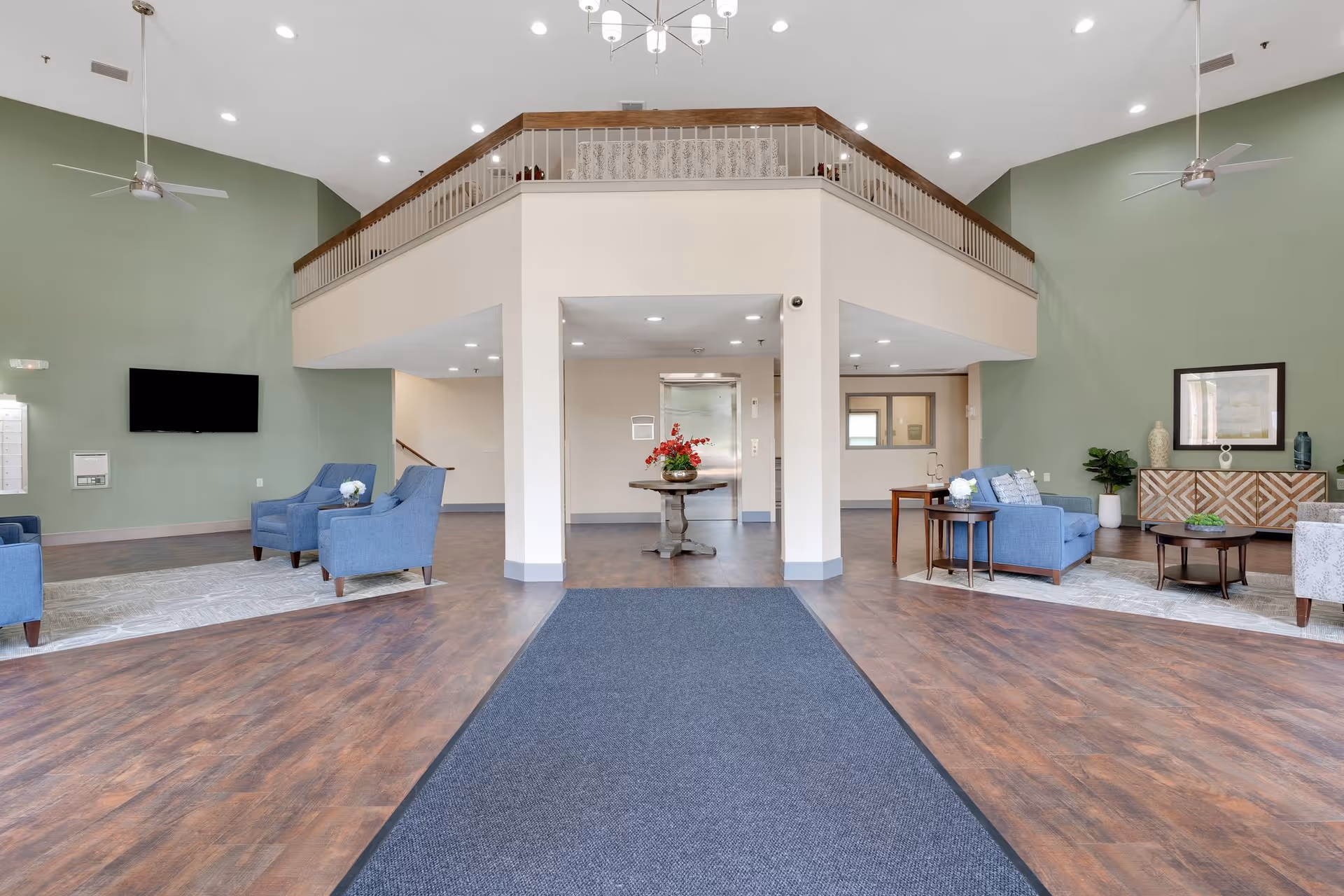 Spacious senior living facility common area with high ceilings, green walls, ceiling fans, and wood flooring. The room features multiple seating areas with blue and gray armchairs and sofas arranged around coffee tables and side tables. A flat-screen TV is mounted on the left wall, and a round table with a flower arrangement is centered near an elevator. A balcony railing overlooks the space from above.