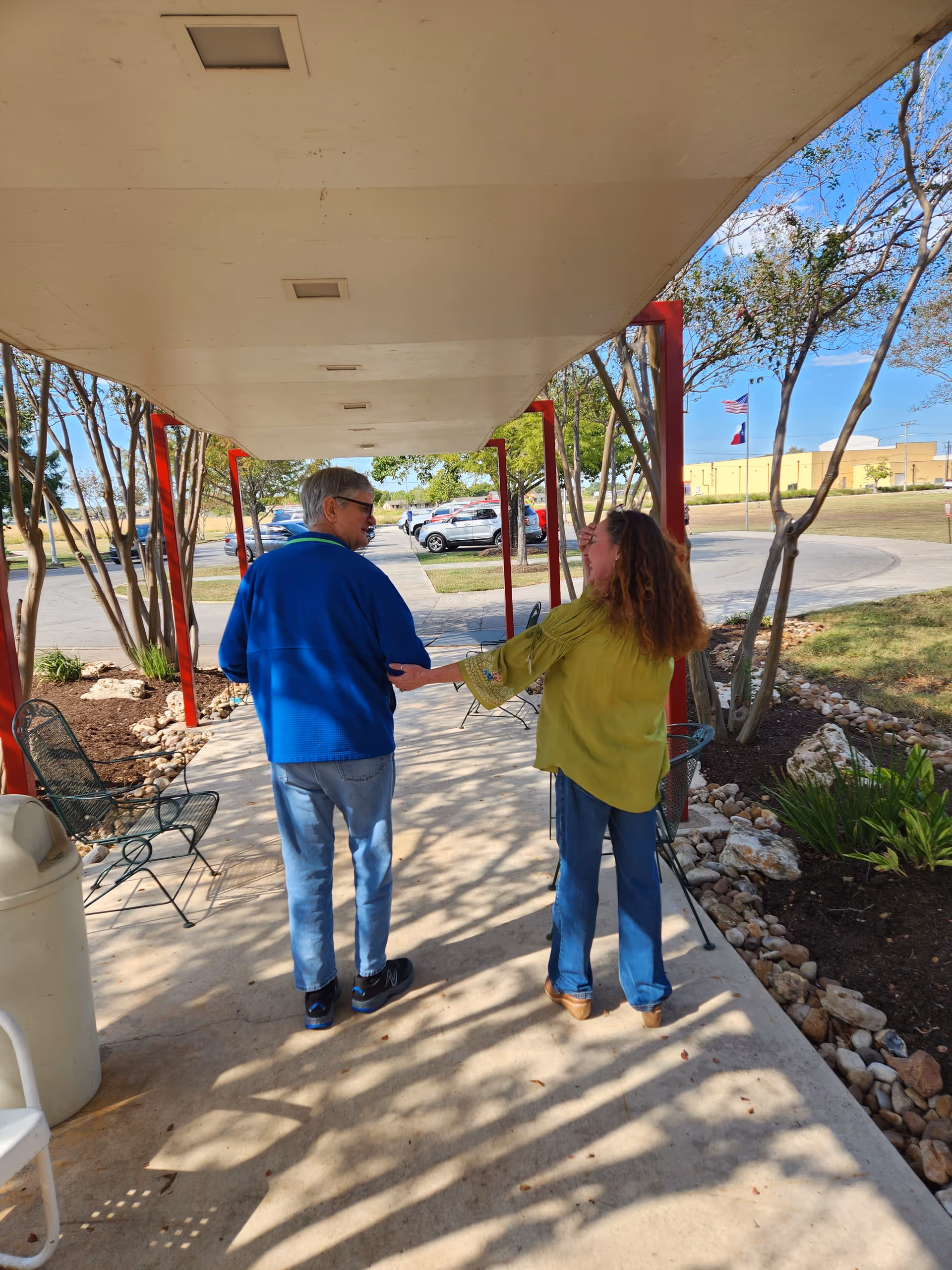 An elderly man and a woman walking together under a covered walkway outside. The man is wearing a blue jacket and jeans, and the woman is wearing a green blouse and jeans. There are trees, chairs, and parked cars visible in the background under a clear blue sky.