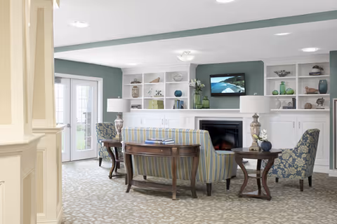 A cozy living room area with a striped sofa and two patterned armchairs arranged around a fireplace with a TV mounted above it. The room has built-in white shelves with decorative items, two side tables with lamps, and a glass door leading outside. The walls are painted green and the floor is carpeted with a light patterned design.