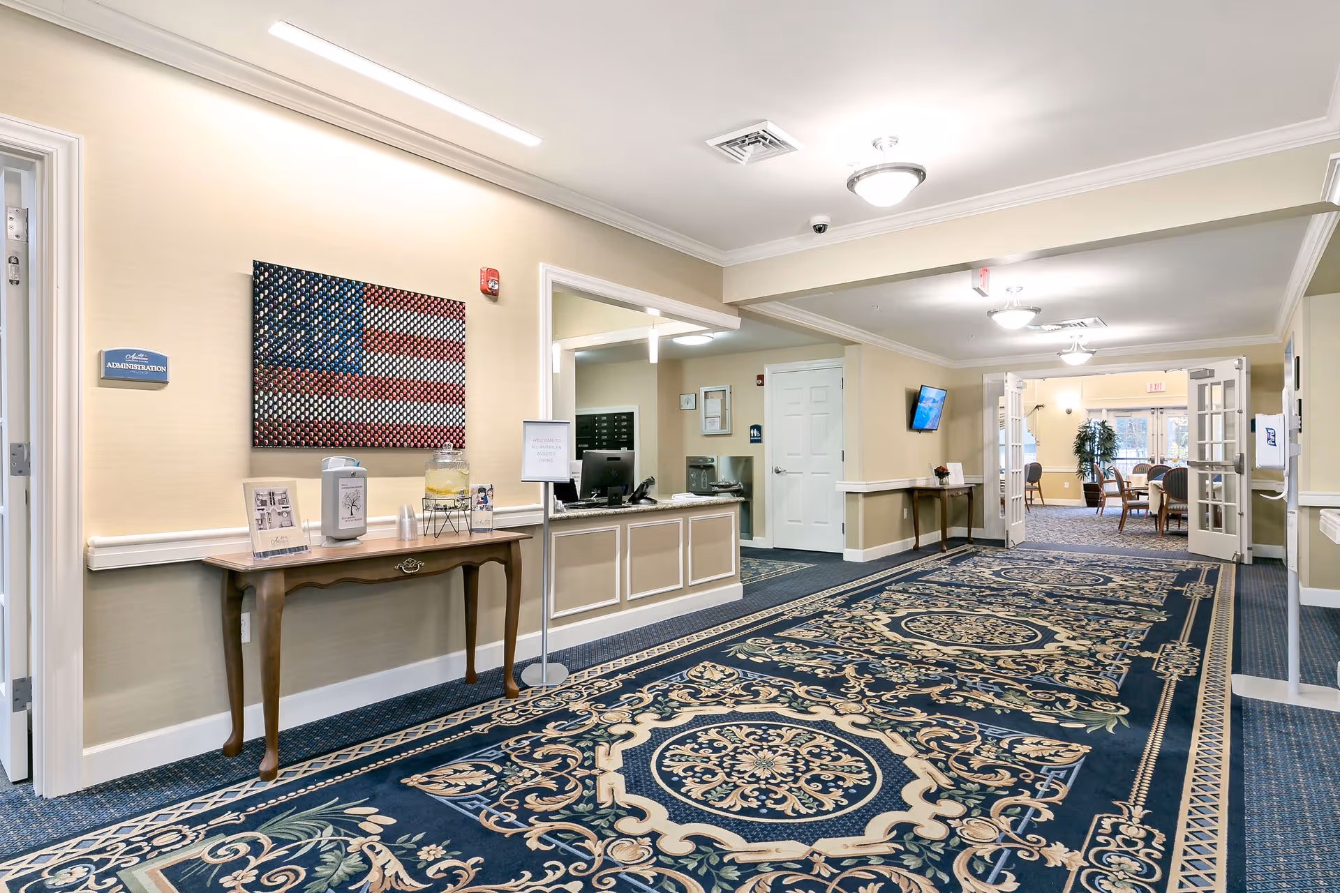 A bright and spacious hallway in a senior living facility with a patterned blue carpet and beige walls. On the left side, there is a wooden table with informational brochures, a water dispenser with cups, and a large wall art piece depicting an American flag made of circular elements. A sign on the wall reads 'Administration.' The hallway leads to a room with tables and chairs visible through open double doors. Ceiling lights illuminate the area.