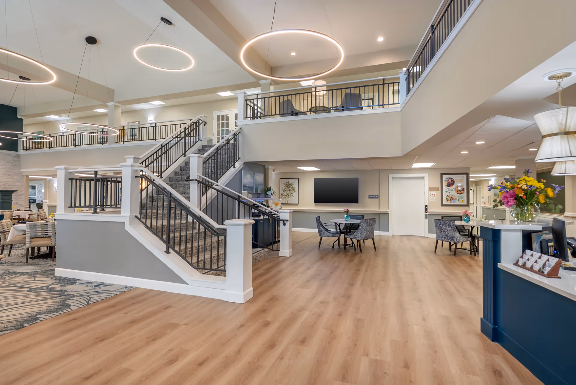 Spacious and modern senior living facility interior with a wide wooden floor area, a staircase leading to an upper level with railings, round tables with chairs, a large flat-screen TV mounted on the wall, and a reception desk with a vase of colorful flowers. Circular pendant lights hang from the ceiling, and the area is well-lit with natural and artificial light.