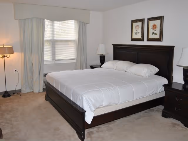 A bedroom with a large dark wooden bed frame and white bedding, two pillows, two matching dark wooden nightstands with lamps, beige carpet, a window with closed blinds and light-colored curtains, and two framed floral pictures hanging above the bed.
