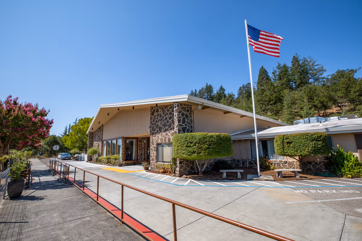 Exterior view of Cloverdale Healthcare Center building with stone and beige walls, a flagpole with an American flag, trimmed bushes, and a clear blue sky. There is a sidewalk with a handrail and some potted plants along the left side.