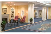 Carpeted senior living common area with a fireplace, upholstered armchairs and a small table near decorative columns and a hallway.