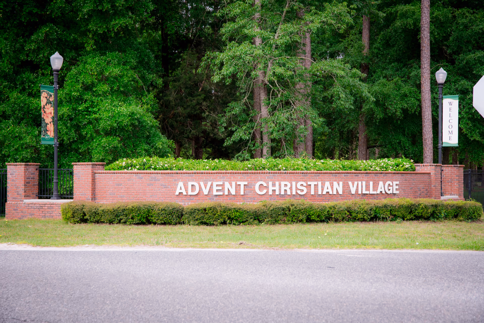 A brick sign with white letters reading 'ADVENT CHRISTIAN VILLAGE' surrounded by green bushes and trees, with two lamp posts on either side, one with a floral banner and the other with a welcome banner.