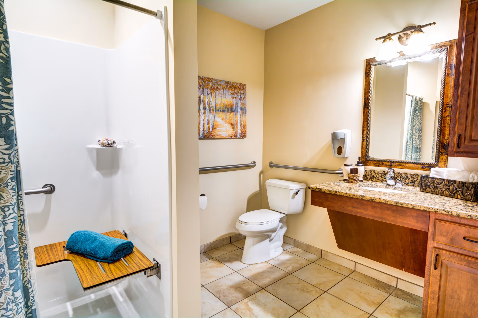 A clean and accessible bathroom featuring a white toilet with grab bars on the walls, a shower area with a fold-down wooden seat and a blue towel placed on it, a granite countertop with a sink, a large framed mirror above the sink, a wall-mounted soap dispenser, and a decorative painting on the wall.