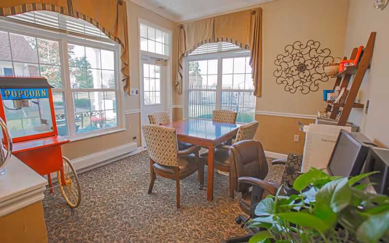 A cozy interior room with a square wooden table surrounded by four upholstered chairs. Large windows with beige curtains let in natural light. A red popcorn machine is positioned near the windows. There is a decorative metal wall art on the right wall and a wooden shelf holding various items. A computer monitor and office chair are visible in the foreground next to a green leafy plant.
