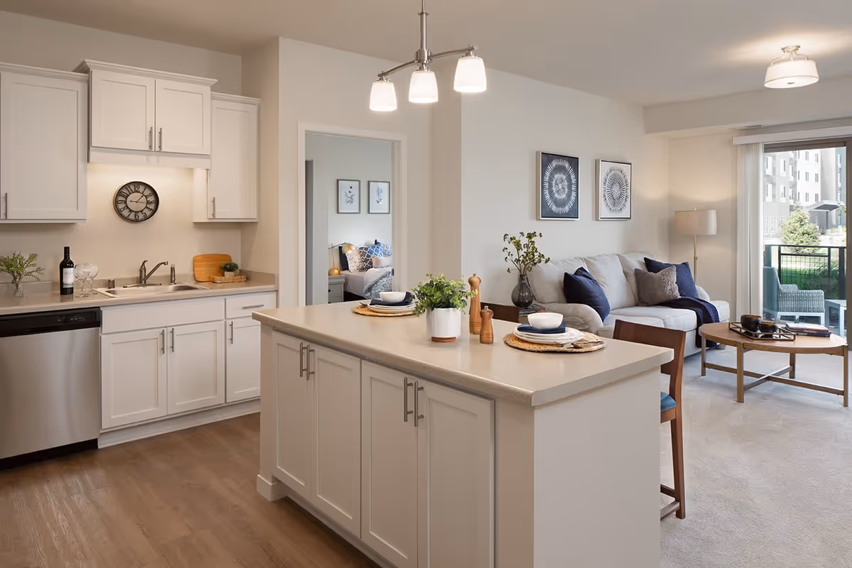 Bright and modern open-concept living space featuring a kitchen with white cabinets, stainless steel dishwasher, and a large island with place settings and potted plants. Adjacent living room has a light gray sofa with blue and gray cushions, a wooden coffee table, floor lamp, and sliding glass door leading to an outdoor patio with seating. A bedroom is visible through an open doorway.