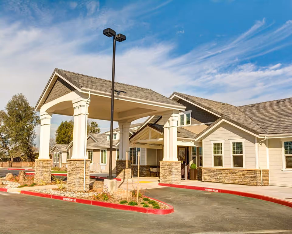 Front entrance of a single-story senior living facility featuring a covered porte-cochère with stone columns, multiple windows, and a driveway.