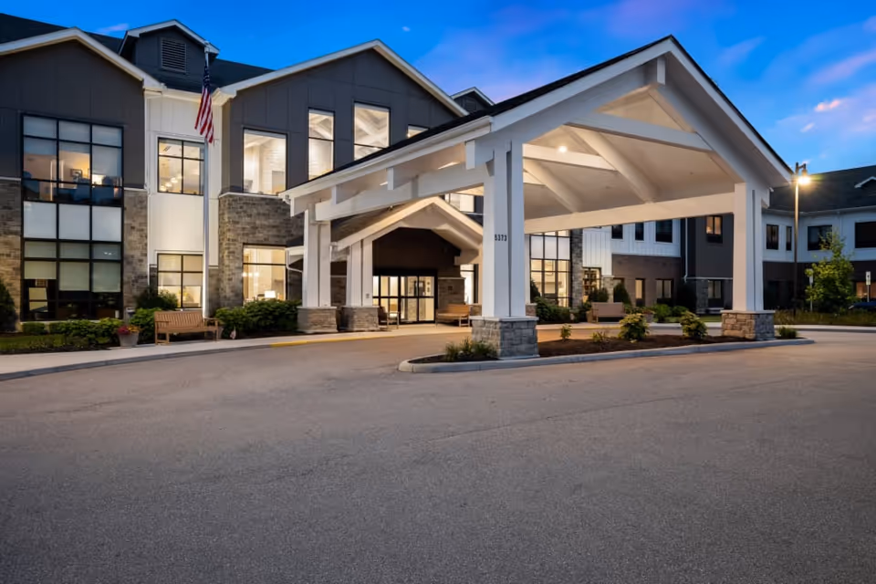 Exterior view of a senior living facility named Montage Mason during dusk. The building features a large covered entrance with white beams and stone pillars, multiple windows, and an American flag mounted near the entrance. There are benches and landscaped greenery around the entrance area.