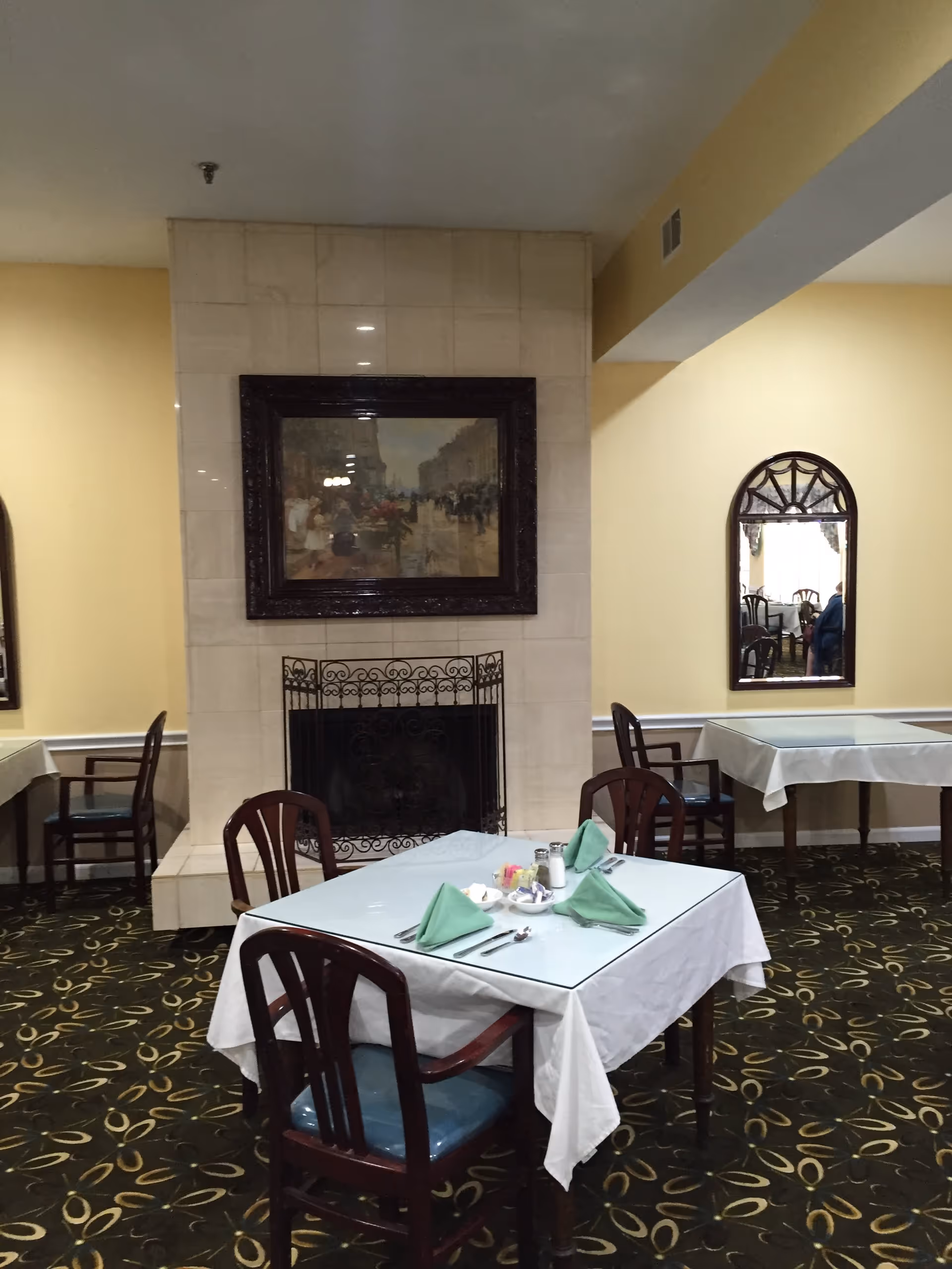 Dining room with tables covered in white tablecloths and green folded napkins, wooden chairs with blue cushions, a fireplace with a decorative screen, a framed painting above the fireplace, and two mirrors on the yellow walls reflecting parts of the room.