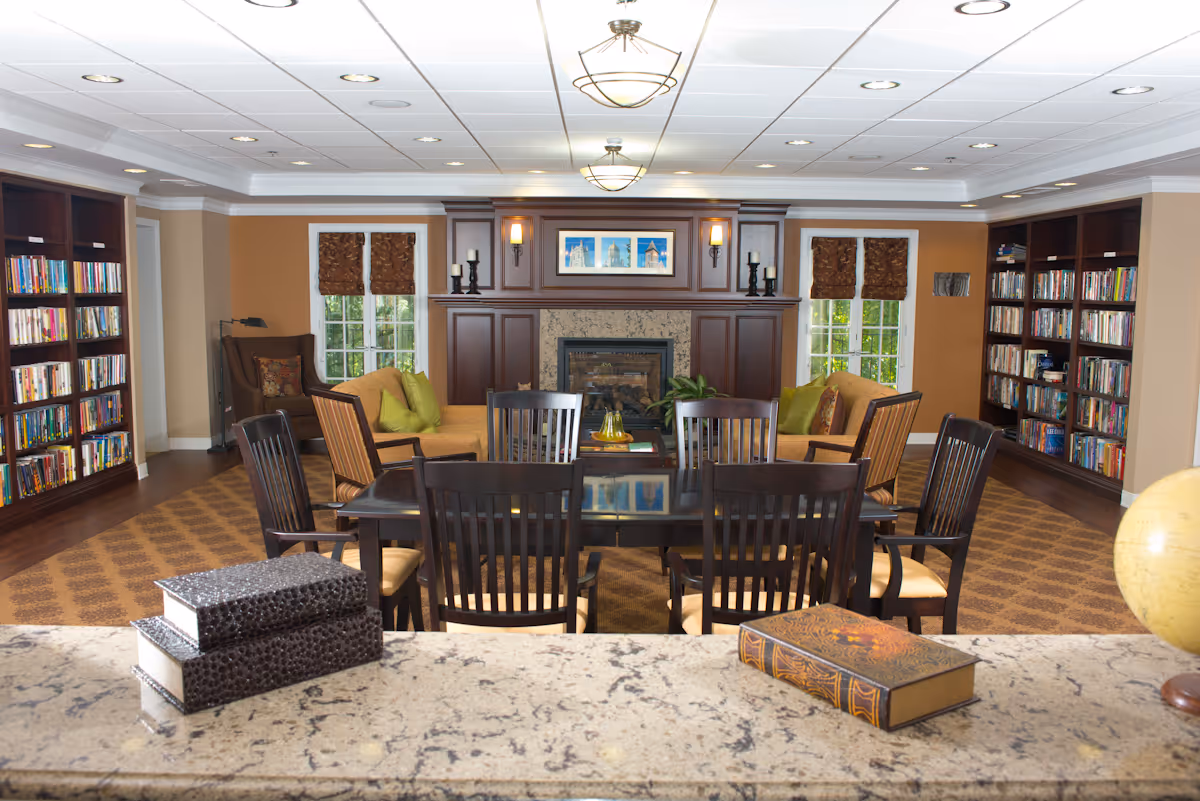 A cozy common area with a fireplace centered on the back wall, flanked by two windows with brown roman shades. There are bookshelves filled with books on both sides of the room. In front of the fireplace, there are two sofas with green and patterned cushions and several wooden chairs around a dark wooden table. The ceiling has recessed lighting and two hanging light fixtures. In the foreground, a granite countertop holds two decorative boxes and a large book, with a globe partially visible on the right.