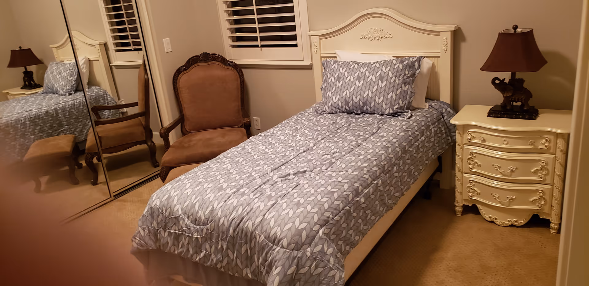 A bedroom with a single bed covered in a blue and white patterned comforter and pillow. Next to the bed is a cream-colored nightstand with ornate detailing and a lamp with a brown shade and an elephant-shaped base. There is a brown upholstered armchair beside the bed and a mirrored closet door reflecting part of the room. The walls are beige and there is a window with white shutters above the chair.