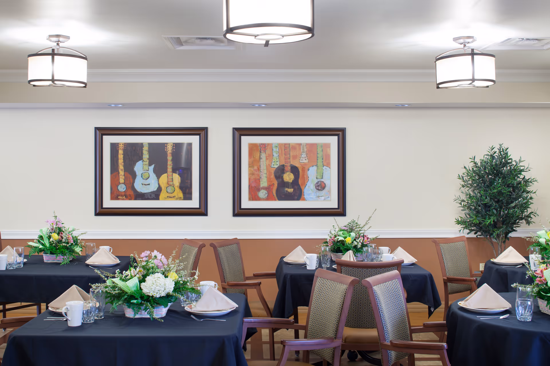 A dining room with tables covered in black tablecloths, each set with beige napkins, white mugs, glasses, and floral centerpieces. The walls are decorated with framed paintings of guitars, and there is a potted plant in the corner. The room is well-lit with ceiling lights.