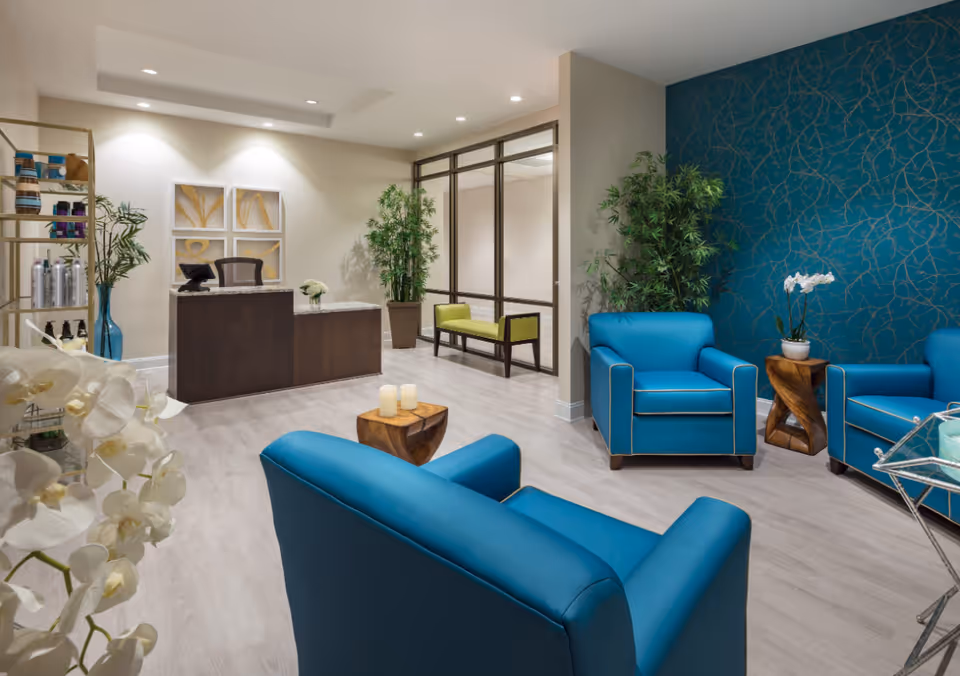 A modern reception area with a dark wooden desk, blue armchairs, a small wooden side table with candles, and decorative plants. The wall behind the armchairs features a blue wallpaper with a gold vine pattern. The floor is light-colored wood, and there is a glass partition with a green bench visible in the background.