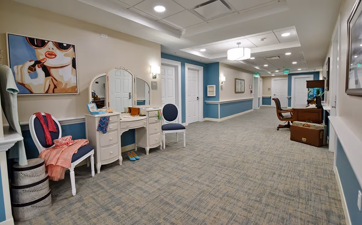 A long, well-lit hallway in a senior living facility with blue and beige walls and carpeted floor. On the left side, there is a white vanity table with a mirror, a chair with clothes draped over it, and a colorful painting of a woman applying lipstick. On the right side, there is a wooden desk with a chair and a small fish tank on top, along with some suitcases on the floor. Several white doors line the hallway, and ceiling lights illuminate the space.