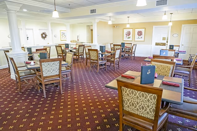 Bright communal dining room with several small tables, upholstered chairs, and menus on each table.