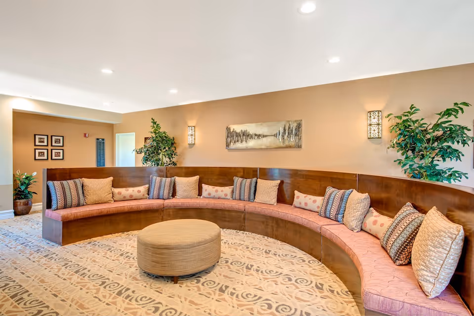 Curved built-in seating with patterned cushions and a round ottoman in a warmly lit lounge area with plants and wall art.
