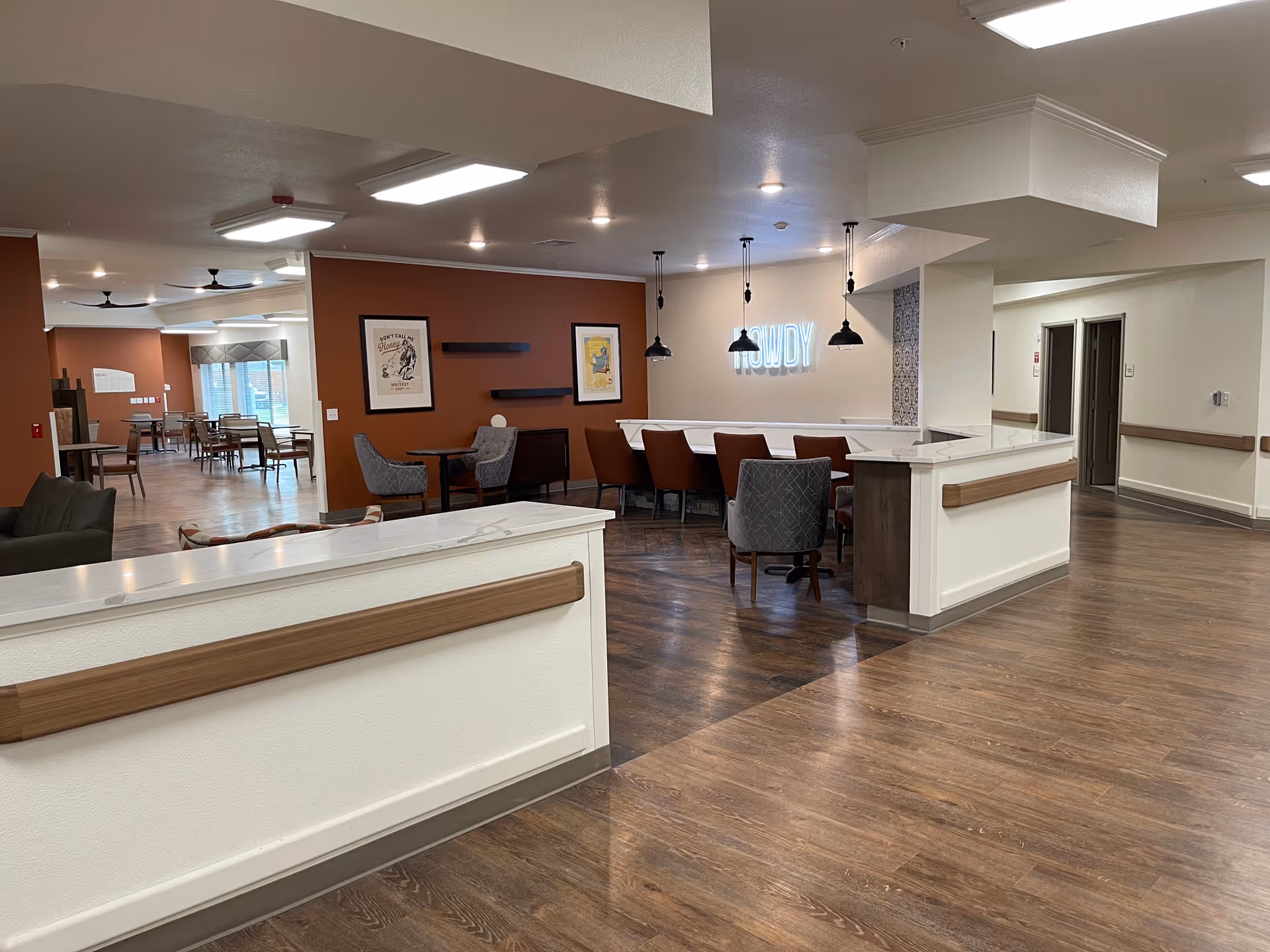 Interior view of a senior living facility common area with wood flooring, white walls, and brown accent walls. There are several seating areas including a bar with high chairs, small tables with chairs, and a lounge area with armchairs. Pendant lights hang above the bar area and a neon sign on the wall reads 'HOWDY'. The space is well-lit with ceiling lights and has a modern, welcoming atmosphere.