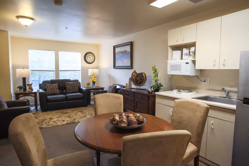 A cozy living area and kitchenette in a senior living facility. The space includes a round wooden dining table with four beige upholstered chairs, a dark leather sofa with patterned cushions, two side tables with lamps, a wall clock, and a framed painting. The kitchenette features white cabinets, a microwave, a sink, and a stainless steel refrigerator. Decorative items are placed on the sideboard and table.
