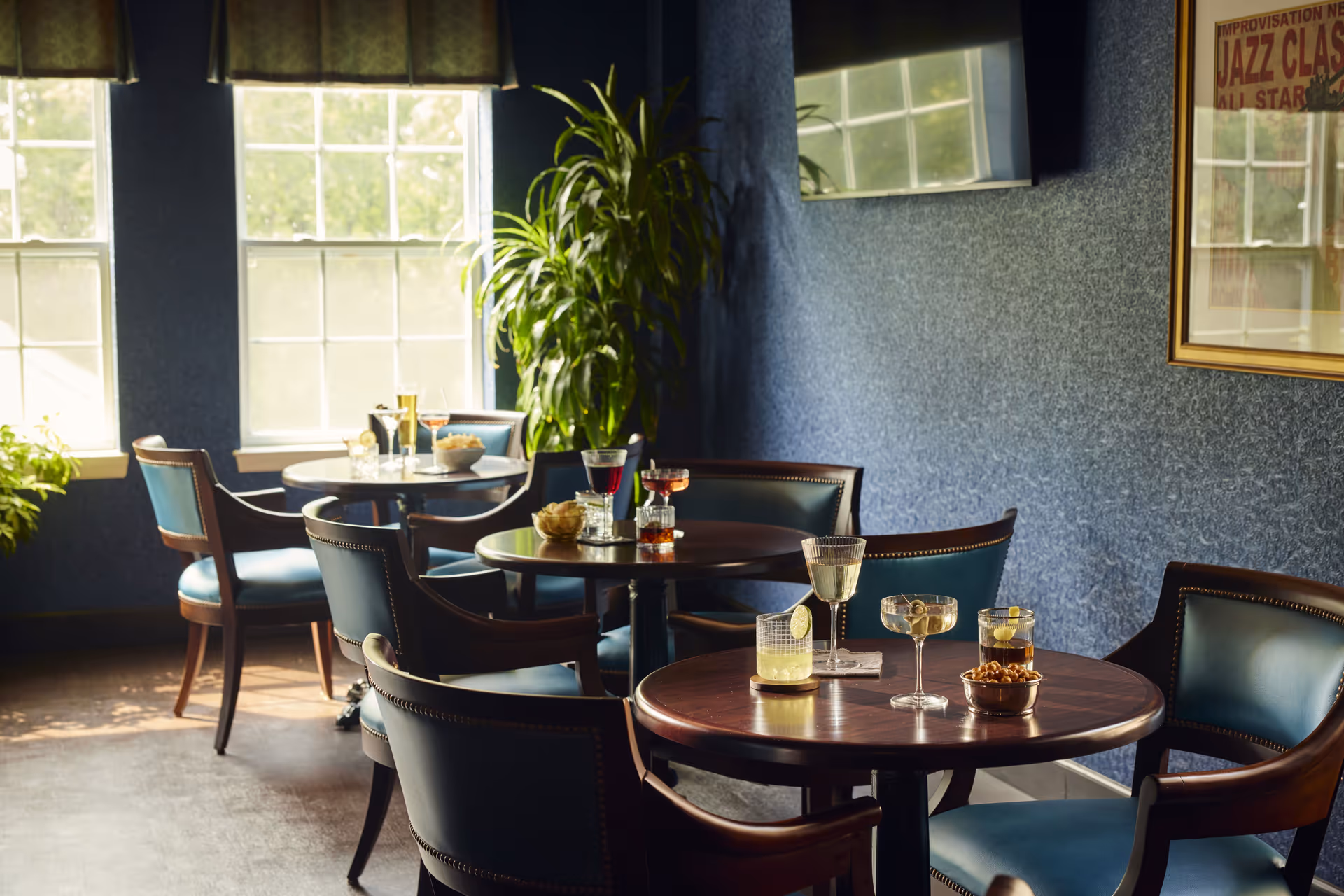 A cozy lounge area with round wooden tables and blue cushioned chairs. Various drinks and snacks are placed on the tables. The room has blue textured walls, two large windows letting in natural light, a large green plant near the window, a wall-mounted TV, and a framed jazz-themed poster on the wall.