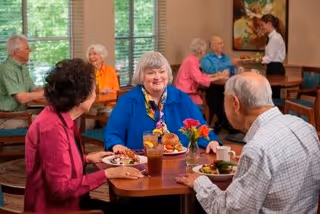A group of elderly people sitting at tables in a dining room, enjoying meals and conversing. A server is attending to a table in the background. The room has large windows with blinds and framed artwork on the walls.