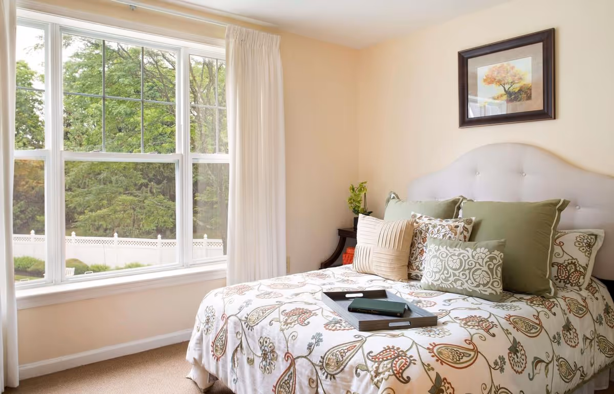 A bright bedroom with a large window showing green trees outside. The bed has a white headboard and is covered with a floral patterned bedspread and multiple decorative pillows in green, beige, and patterned designs. A small dark wooden nightstand with a plant is beside the bed, and a framed picture of a tree hangs on the wall above the headboard.