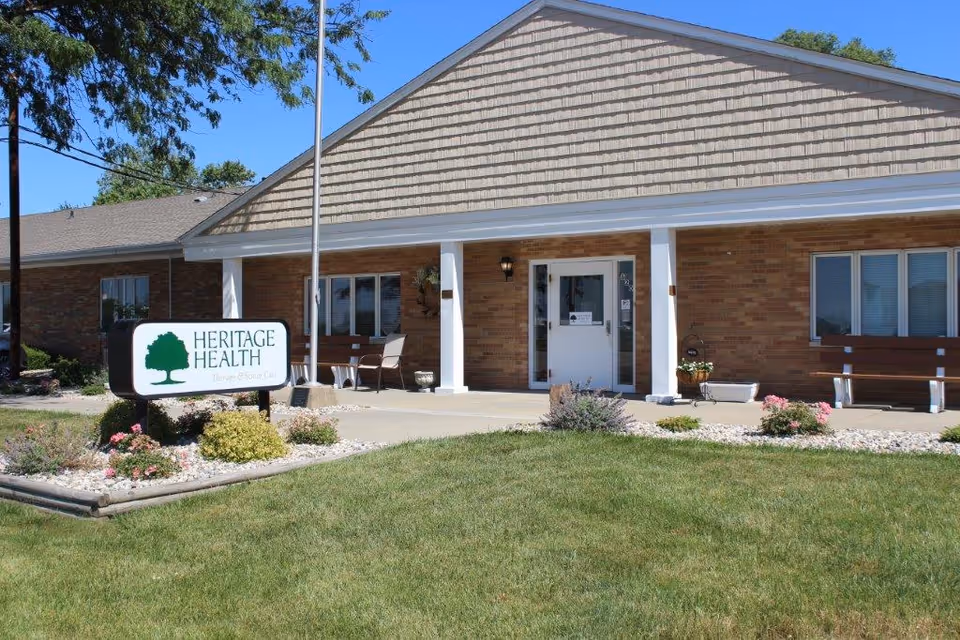 Exterior view of a single-story brick building with a peaked roof and a covered entrance supported by white columns. There are benches and chairs on the porch area. A sign in front of the building reads 'Heritage Health Therapy & Senior Care'. The surrounding area has a well-maintained lawn, flower beds, and a flagpole.