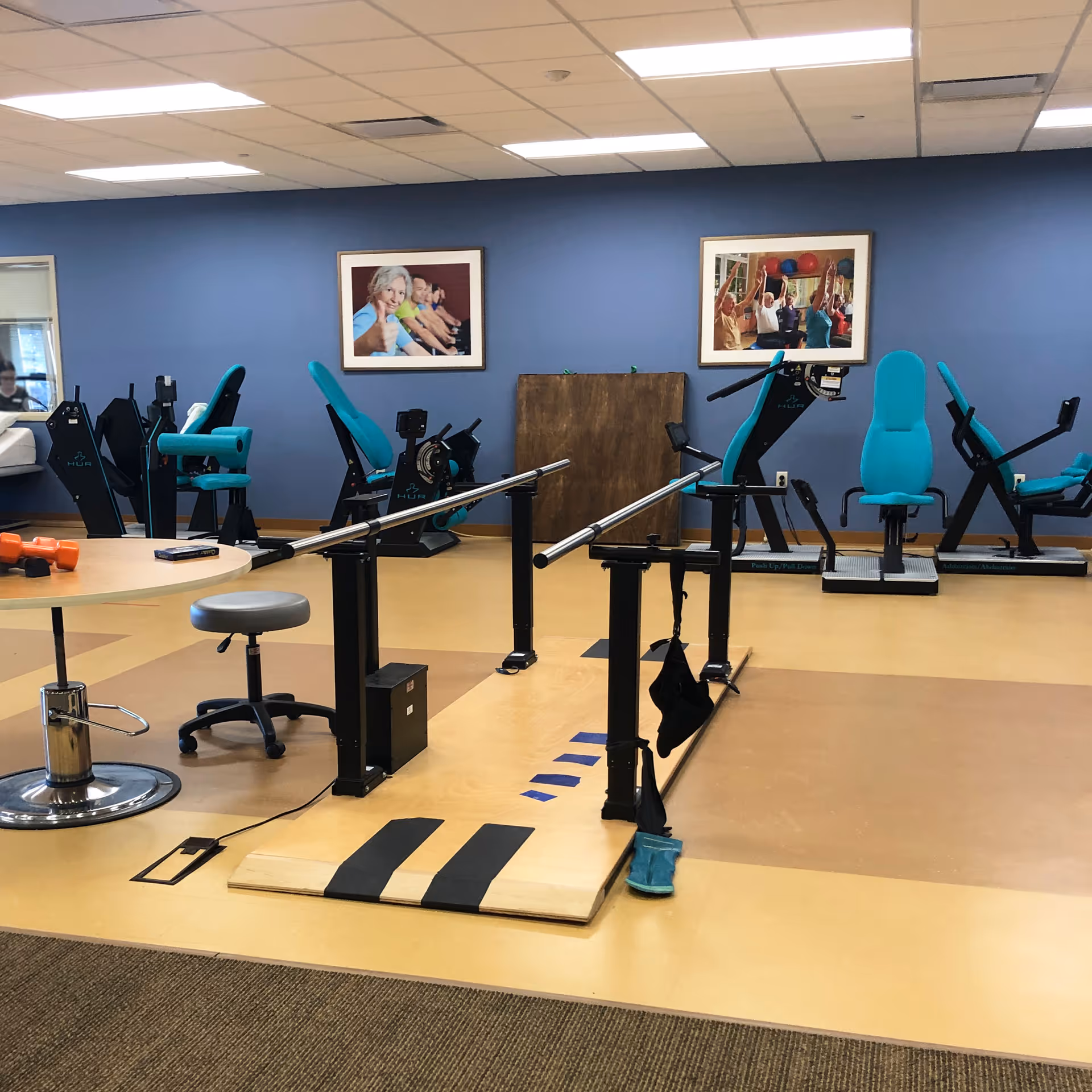 Indoor physical therapy/exercise room with parallel bars, teal exercise machines, and framed photos on a blue wall.