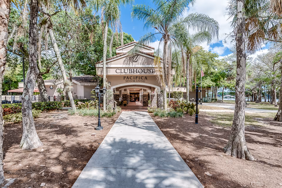 View of the entrance to a senior living clubhouse named Pacifica Senior Living, with a paved walkway leading through a landscaped area with palm trees and other greenery under a partly cloudy blue sky.