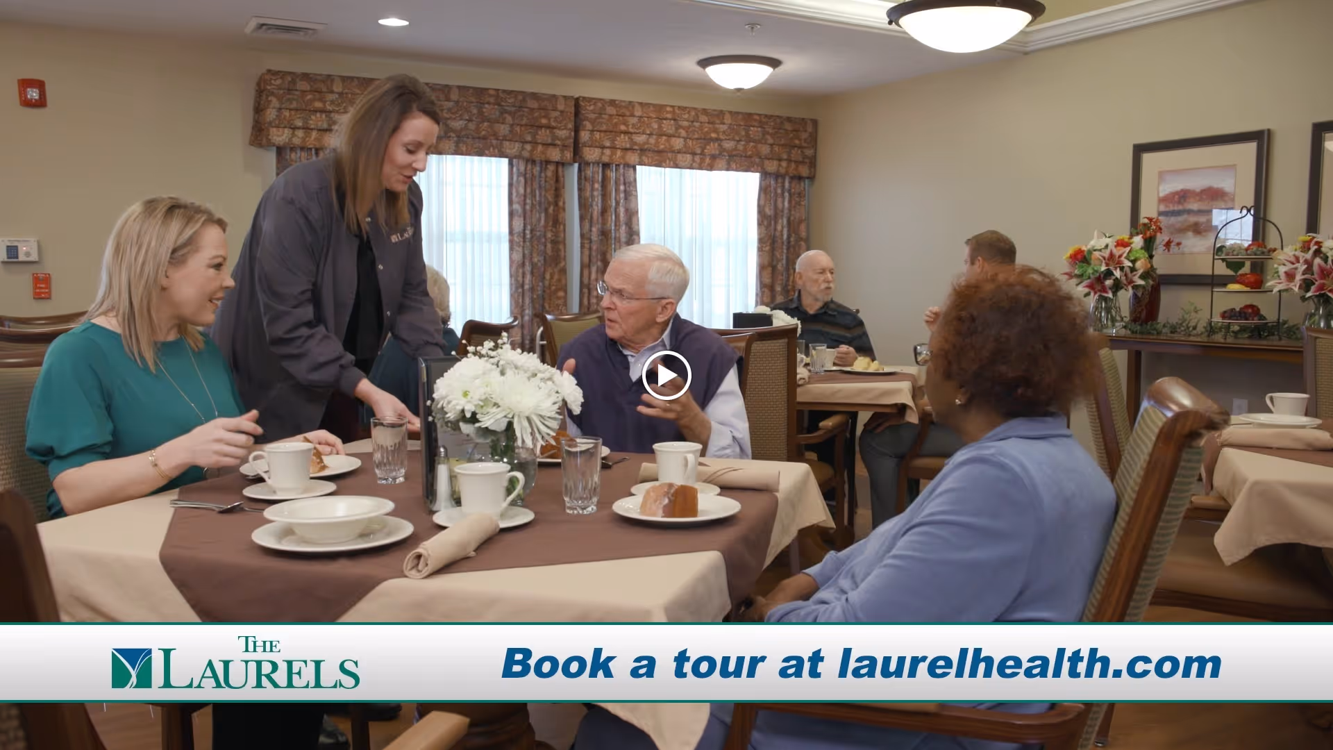 A group of elderly residents and a staff member interacting in a dining room setting at The Laurels of Mt. Vernon. The table is set with plates, cups, and a centerpiece of white flowers. The room has warm lighting, framed artwork on the walls, and floral decorations on a side table.