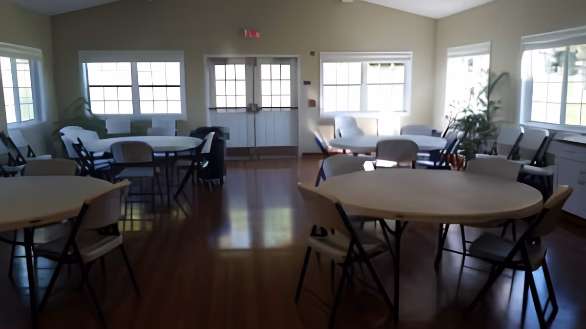 A bright room with multiple round tables and chairs arranged around them, large windows letting in natural light, and a door at the far end. The room has light-colored walls and a polished wooden floor, with some plants placed near the windows.