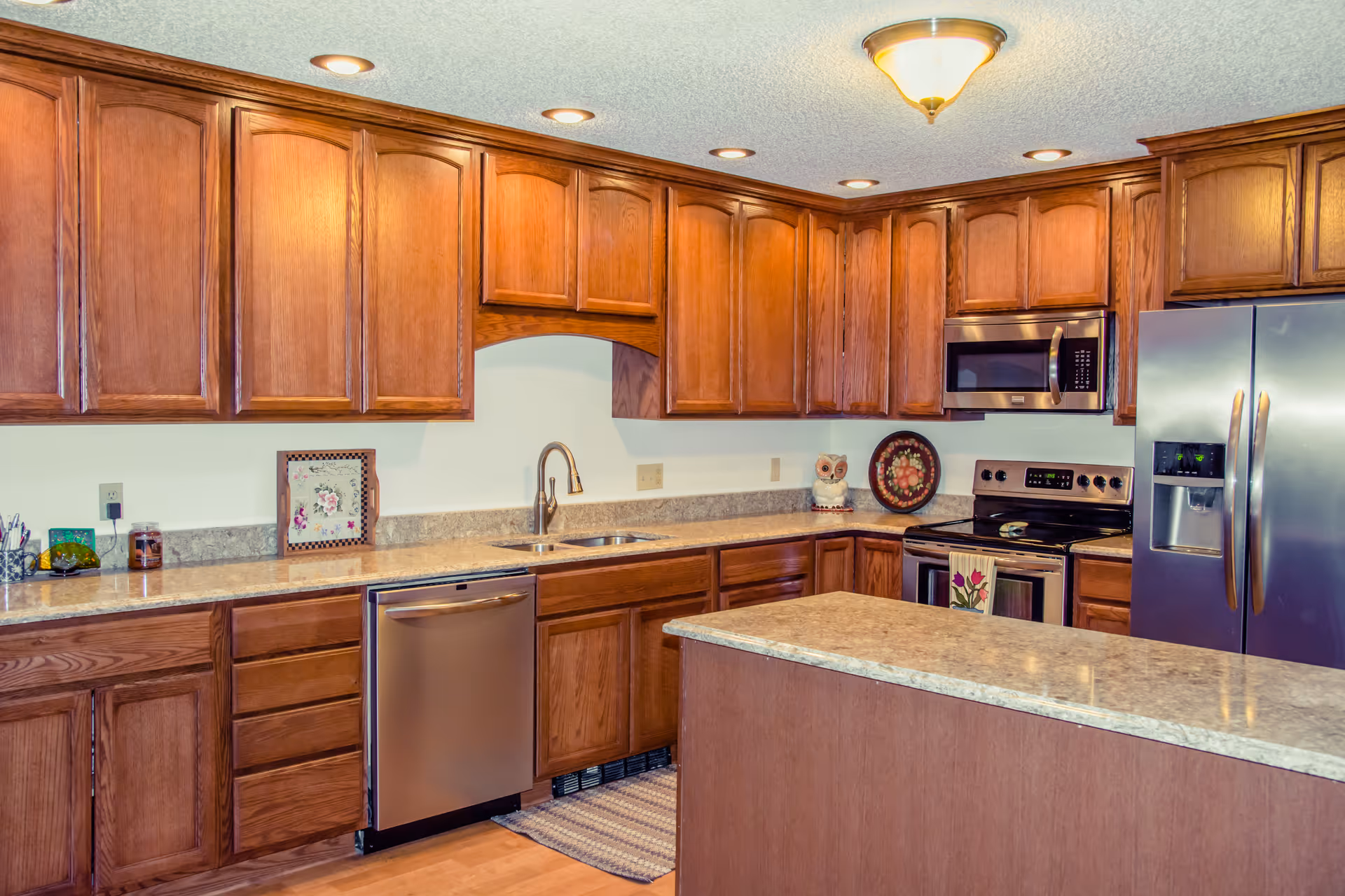 A spacious kitchen with wooden cabinets, a stainless steel refrigerator, dishwasher, microwave, and stove. The kitchen features a large island with a granite countertop, a double sink, and recessed lighting in the ceiling.