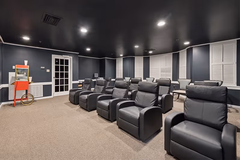 A small theater room with two rows of black leather recliner chairs facing a screen. The room has dark walls and ceiling with recessed lighting, beige carpet, white window shutters, and a red popcorn machine in the corner near a white door with glass panes.