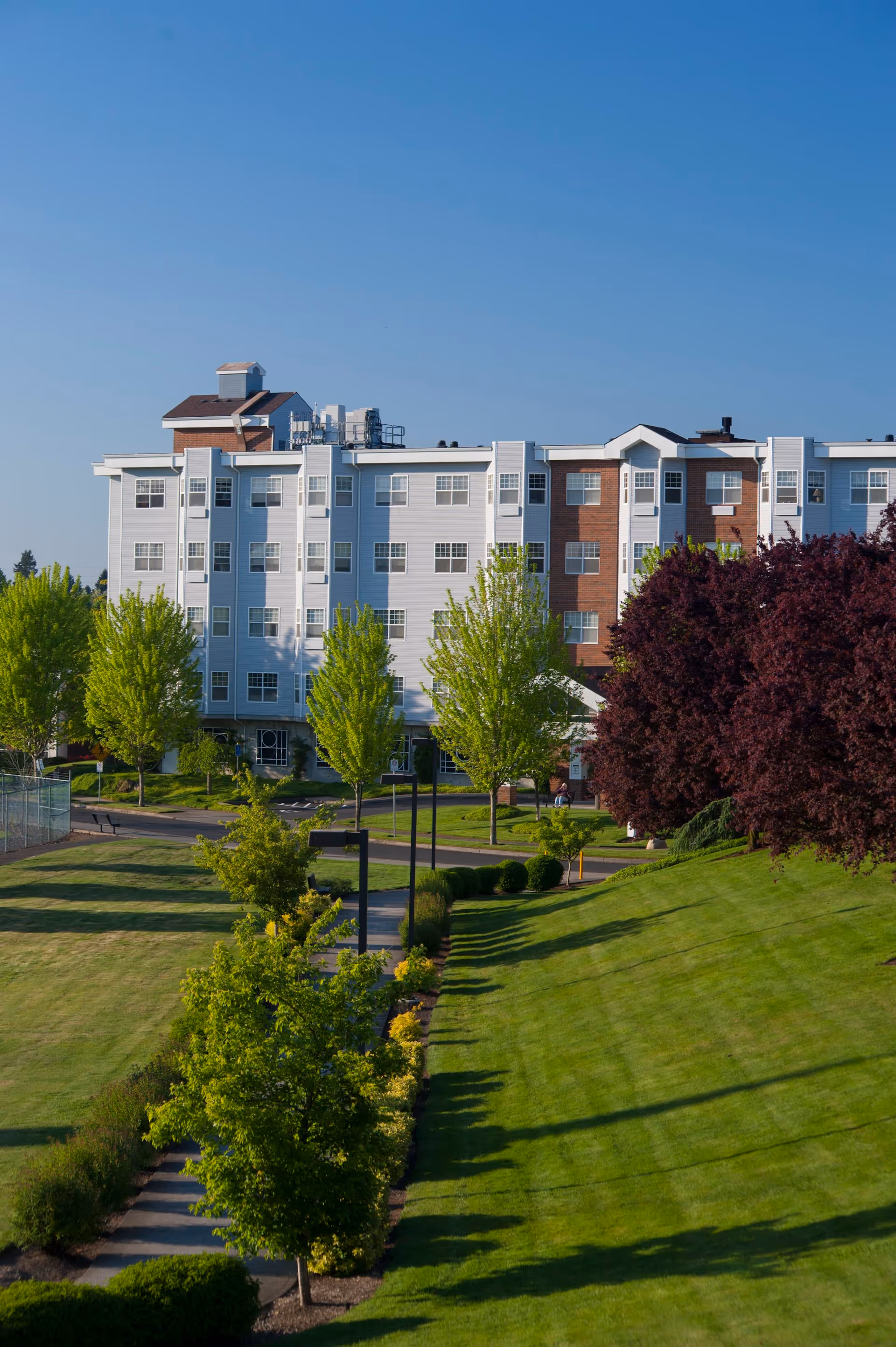 Exterior view of a multi-story senior living facility building with white and brick facade, surrounded by green lawns, trees, and a paved walkway under a clear blue sky.